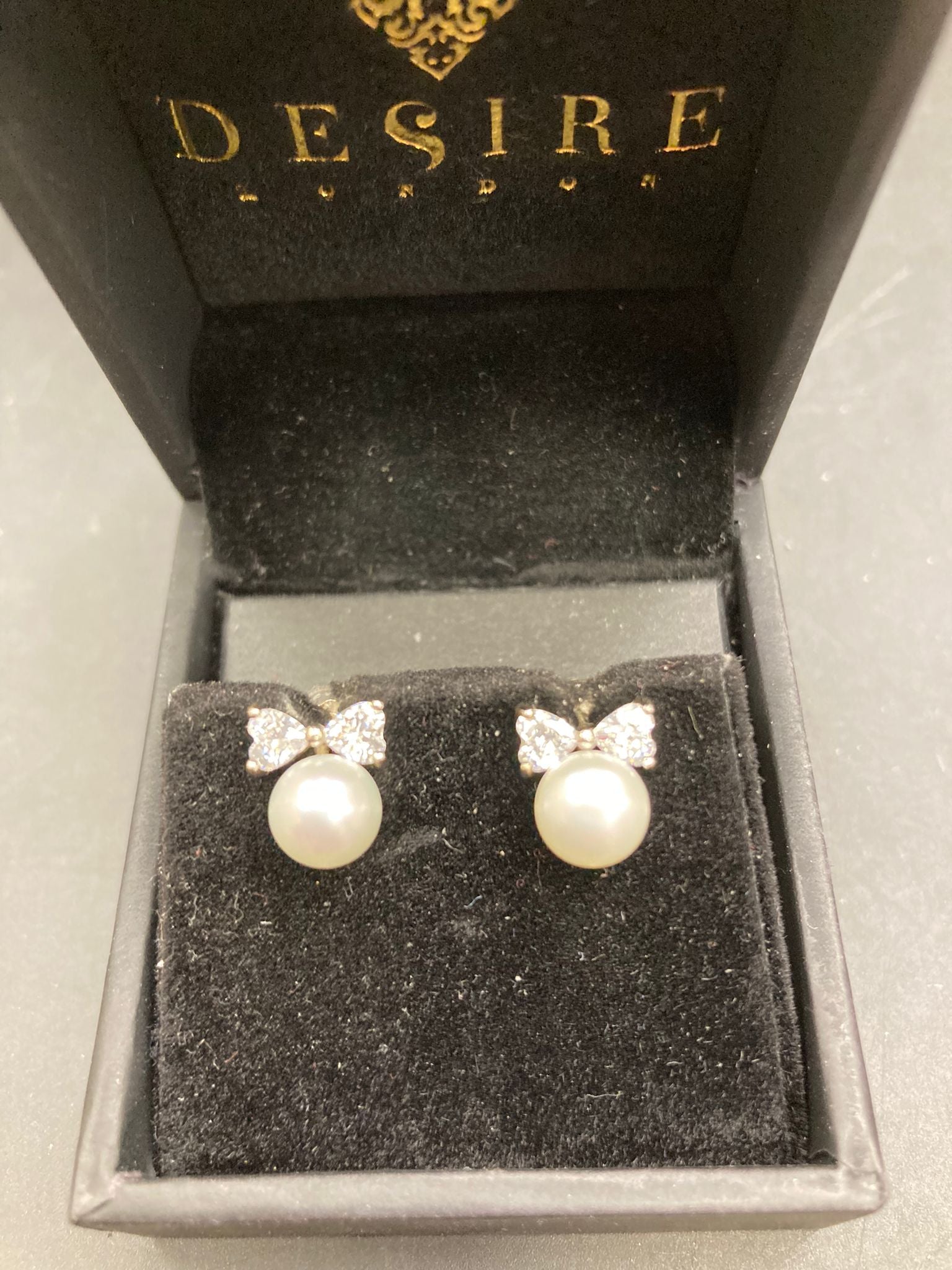 Sterling Silver Cz Bow Pearl Studs in a luxurious Desire London box on a black velvet background.