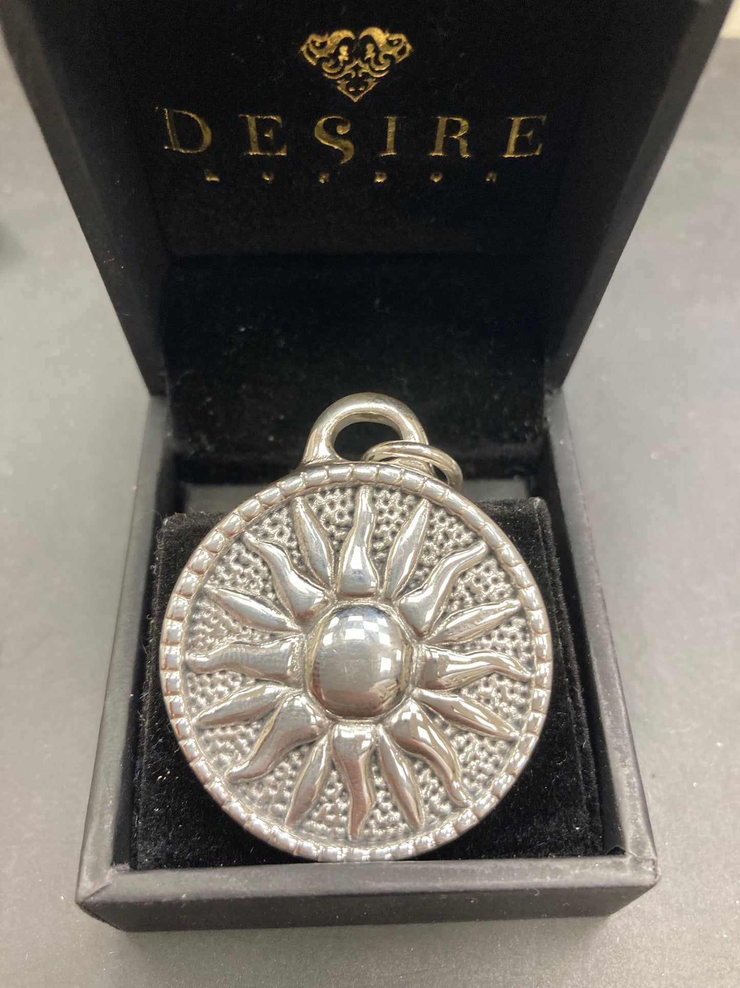 Sterling silver sun pendant displayed in a black box with the logo DE$IRE, showcasing intricate sun design.