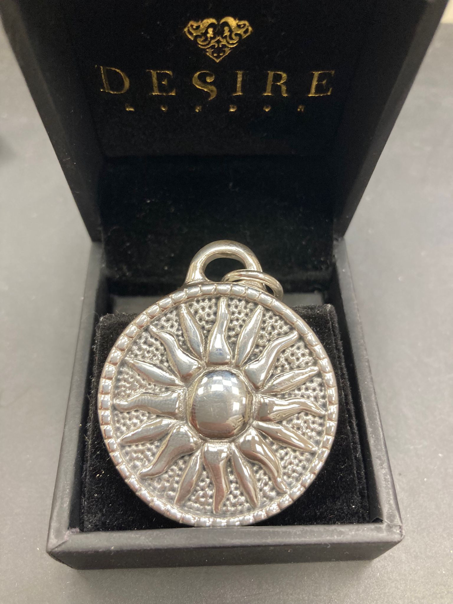 Sterling silver sun pendant displayed in a black box with the logo DE$IRE, showcasing intricate sun design.